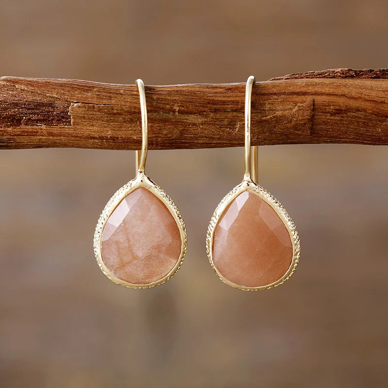A pair of gold-colored sunstone earrings hanging from a wooden branch, with a textured surface and a teardrop shape. | ALLORA JADE
