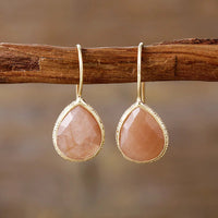 A pair of gold-colored sunstone earrings hanging from a wooden branch, with a textured surface and a teardrop shape. | ALLORA JADE