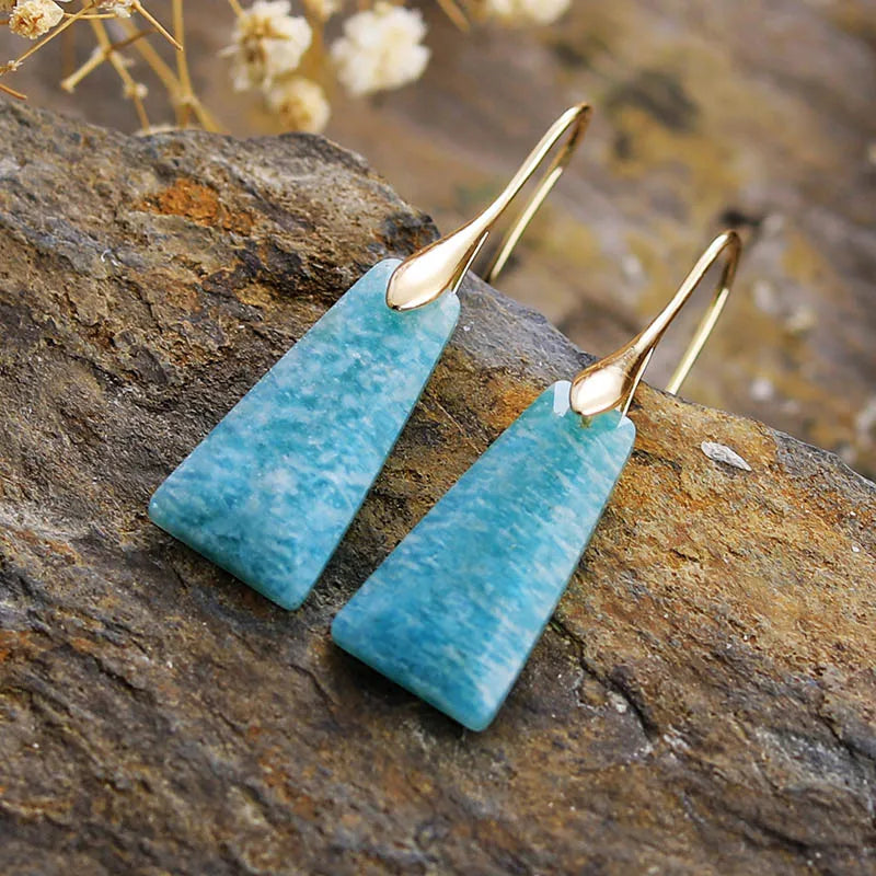A pair of gold-colored brass earrings with blue Amazonite stones, displayed on a rock. ALLORA JADE
