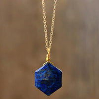 A hexagonal Lapis Lazuli pendant necklace with a gold-coloured chain, displayed against a wooden background. ALLORA JADE