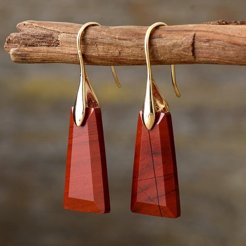 A pair of gold-colored brass drop earrings with red jasper stones, displayed hanging on a wooden branch. ALLORA JADE
