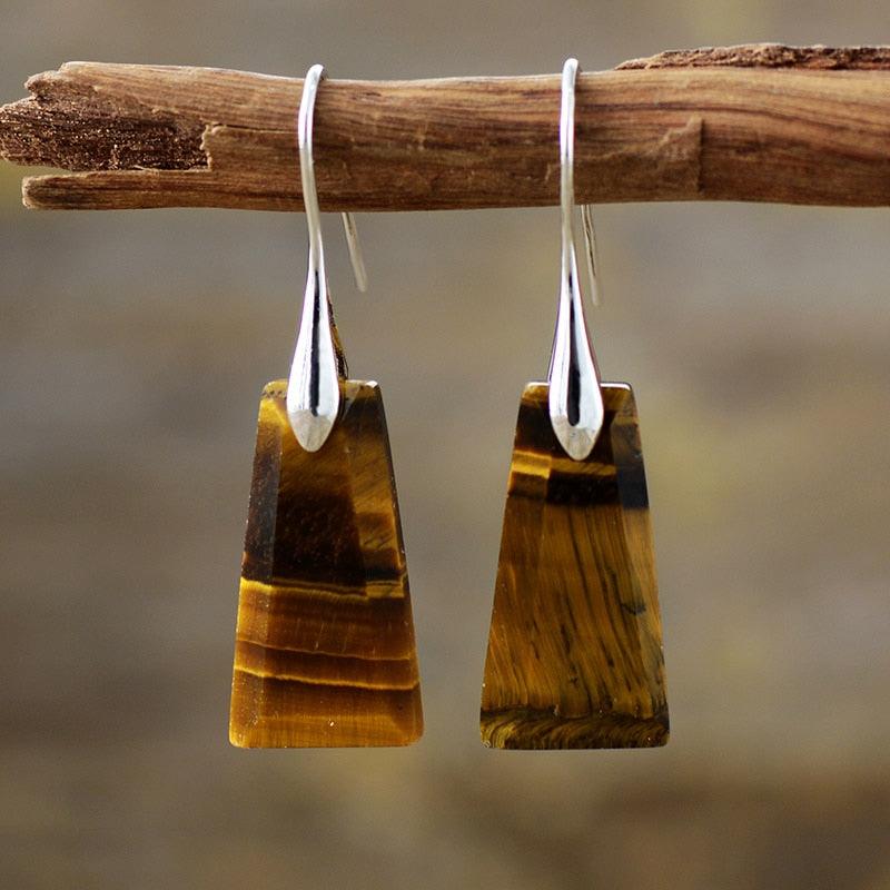 A pair of silver-coloured Tiger's Eye Ascend earrings dangle from a wooden branch against a blurred background. | ALLORA JADE