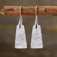 A pair of rose quartz stone earrings with silver-coloured brass, hanging from a wooden branch. ALLORA JADE