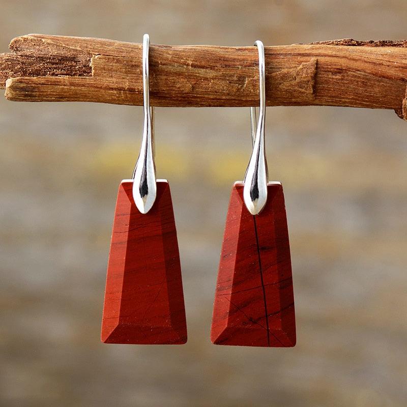 A pair of silver hook earrings with red jasper drop pendants, displayed hanging on a wooden branch. ALLORA JADE