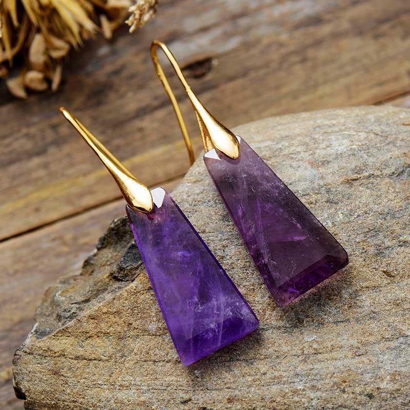 A pair of amethyst earrings with gold-colored brass caps, displayed on a stone surface. | ALLORA JADE
