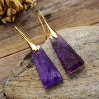 A pair of amethyst earrings with gold-colored brass caps, displayed on a stone surface. | ALLORA JADE