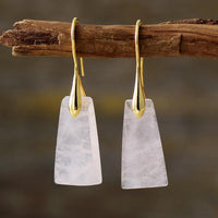 A pair of gold-coloured brass earrings with rose quartz stones, suspended from a branch. ALLORA JADE