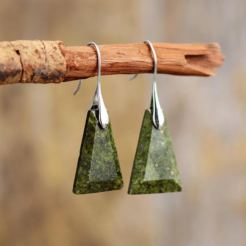 A pair of Green Jasper stone earrings, with a trapezoid shape, suspended from silver-colored brass hooks on a wooden branch. ALLORA JADE