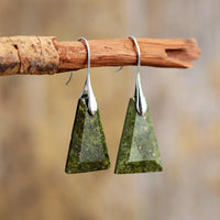 A pair of Green Jasper stone earrings, with a trapezoid shape, suspended from silver-colored brass hooks on a wooden branch. ALLORA JADE