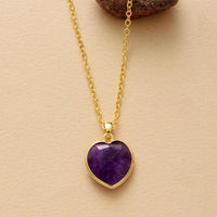 A heart-shaped Amethyst pendant necklace on a gold chain, displayed against a beige background.