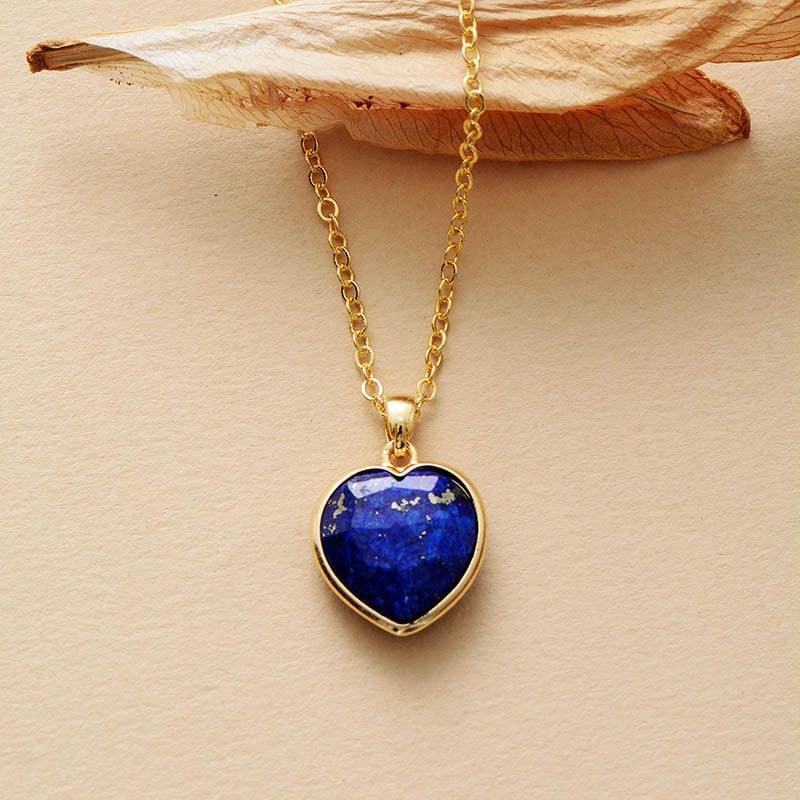 A heart-shaped Lapis Lazuli pendant necklace in a gold-colored setting, displayed against a beige background with a dried leaf. ALLORA JADE