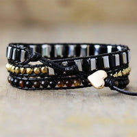 Black leather bracelet with black beads and gold accents, displayed  on a stone surface