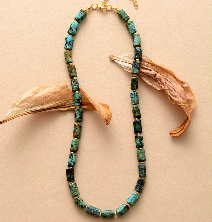 A choker necklace with green African Turquoise beads, displayed against a beige background with some dried plant leaves to the side. ALLORA JADE