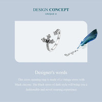 Adjustable silver cross ring with black zircon on a card with a feather and pen, against a light blue background.