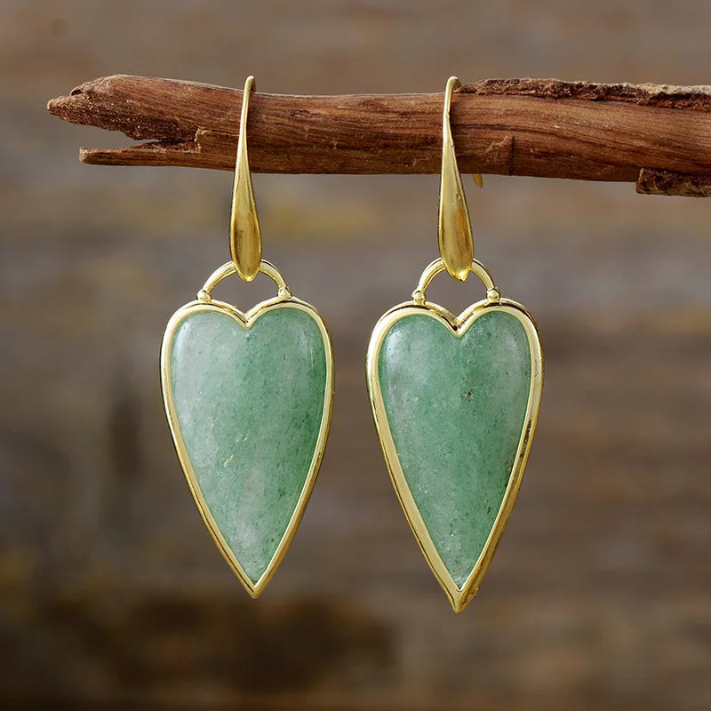 A pair of heart-shaped dangle earrings with polished gold-toned metal borders and hooks, featuring natural green aventurine stones, hanging from a rustic wooden branch against a blurred brown background.