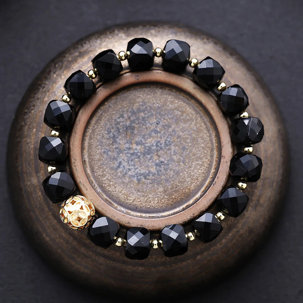 Bracelet with black beads and gold accents on a textured surface. Black Onyx Faceted Cube Bracelet | ALLORA JADE