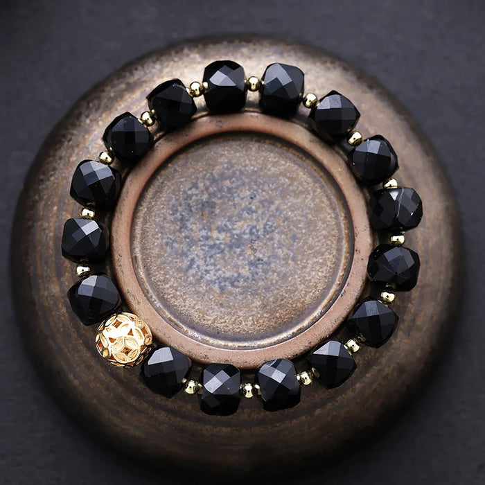 Bracelet with black beads and gold accents on a textured surface. Black Onyx Faceted Cube Bracelet | ALLORA JADE