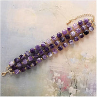 3-strand Purple Amethyst beaded bracelet with gold accents displayed flat on a textured surface.