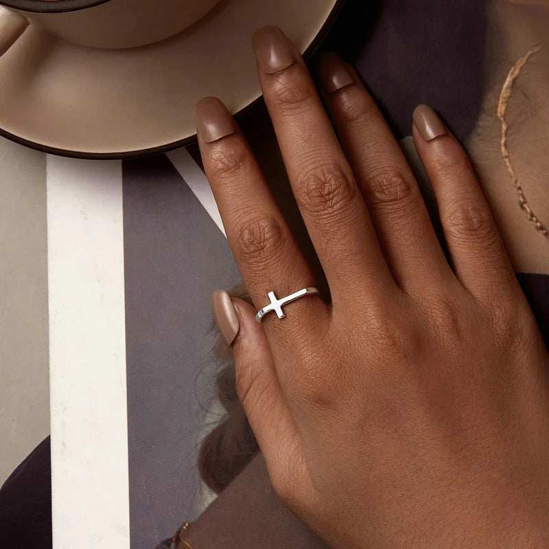 Hand wearing a silver ring with a cross design on a neutral background