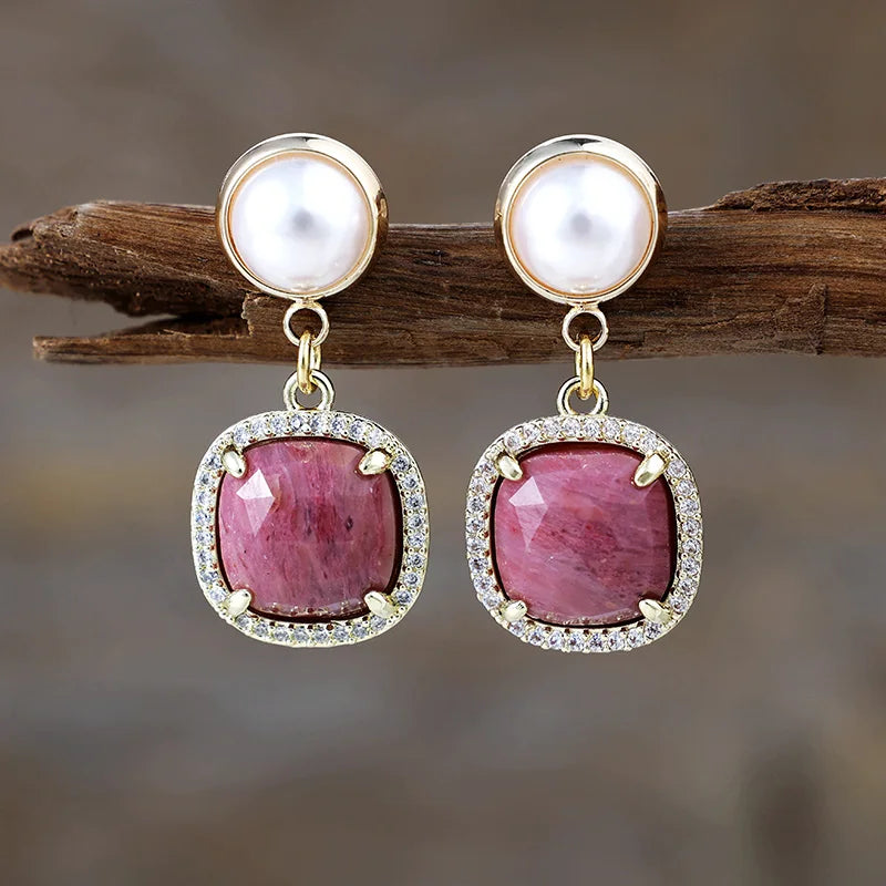 Rhodonite Pavé Cushion Dangle Earrings featuring rosy-pink gemstone drops framed by sparkling rhinestones, with imitation pearl studs in gold-coloured settings, displayed on rustic wood.