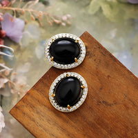 Black Onyx Pavé Oval Stud Earrings featuring polished black gemstones framed by sparkling rhinestones in gold-coloured settings, displayed on rustic wood.