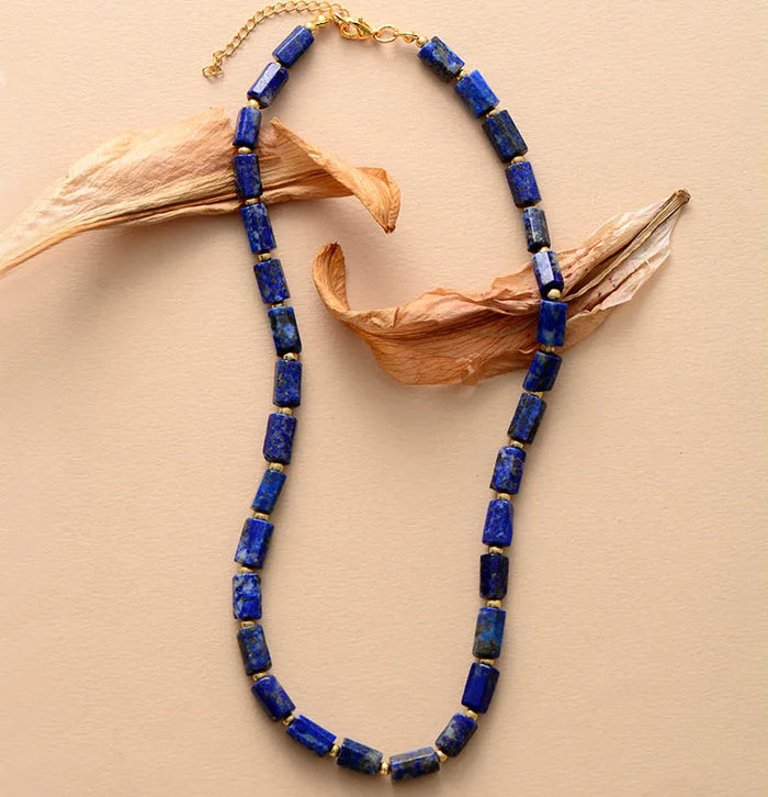 A necklace with rectangular blue Lapis Lazuli beads and gold-colored accents, displayed against a neutral background with a dried leaf. ALLORA JADE