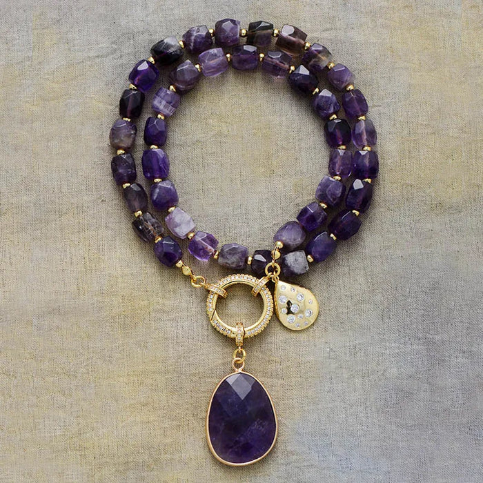 Purple beaded necklace with gold accents and charms on a textured beige background. Amethyst Pavé Statement Necklace | ALLORA JADE