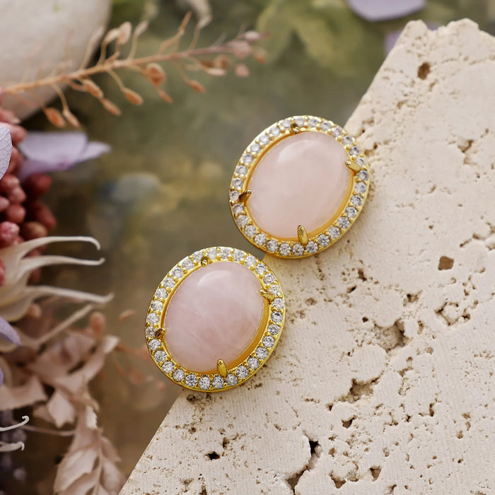 Rose Quartz Pavé Oval Stud Earrings featuring polished pink gemstones framed by sparkling rhinestones in gold-coloured settings, displayed on stone surface with floral background.