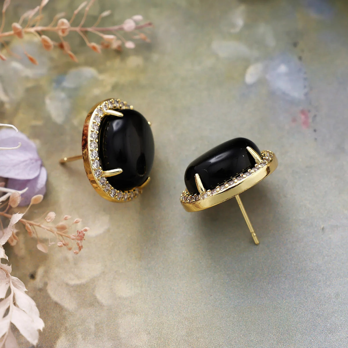 Black Onyx Pavé Oval Stud Earrings featuring polished black gemstones framed by sparkling rhinestones in gold-coloured settings, displayed on floral background.