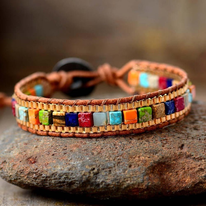 A multicolored Chakra Jasper beaded bracelet with a genuine tan leather cord and an easy-hook clasp, displayed on a stone.