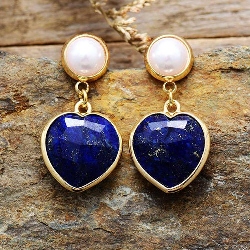 A pair of heart-shaped Lapis Lazuli earrings with gold-colored copper and imitation pearls, displayed on a natural stone background.